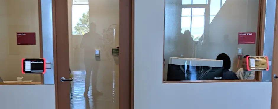 A distant photo showing some conference rooms with glass windows and doors. Next to the doors are some blurry meeting tablets. Also in the glass reflection is a faint image of me!