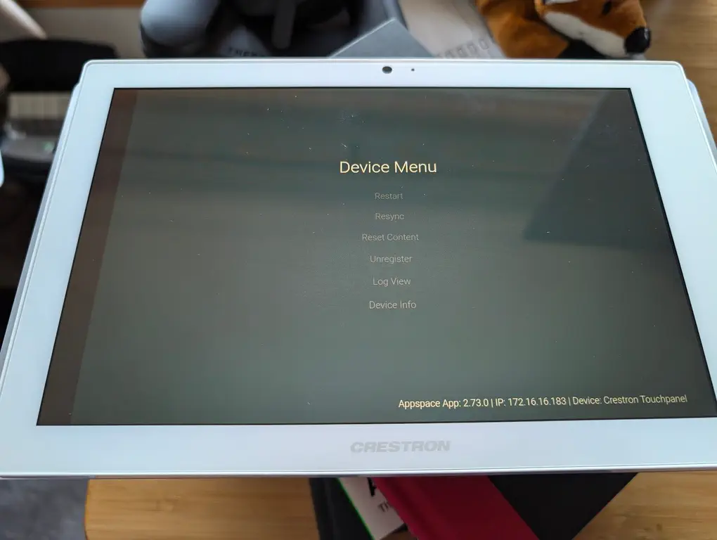 A tablet displays the 'Device Menu' centered with options: Restart, Resync, Reset Content, Unregister, Log View, Device Info. In the bottom right of the screen is a small bit of text saying 'Appspace App: 2.73.0 | IP: 172.16.16.183 | Device: Crestron Touchpanel'