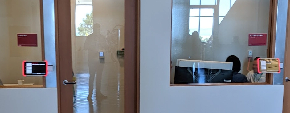 The only photo I have of said cool tablets at Facebook's campus. A distant photo showing some conference rooms with glass windows and doors. Next to the doors are some blurry meeting tablets. Also in the glass reflection is a faint image of me!